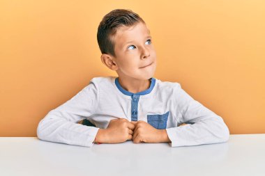 Sevimli beyaz çocuk, günlük kıyafetler giymiş masaya oturmuş gülümsüyor, yan tarafa bakıyor ve düşünüyor.. 