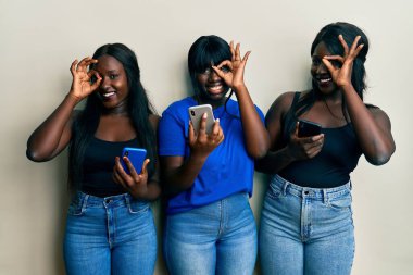 Üç Afro-Amerikalı arkadaş akıllı telefon kullanıyor. El hareketleriyle gülümsüyorlar. Gözleri, mutlu yüzleriyle parmaklarının arasından bakıyor.. 