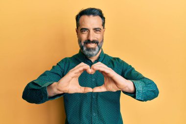 Orta yaşlı, sakallı, gri saçlı, iş kıyafetleri giyen, aşkta gülümseyen, elleriyle kalp sembolü yapan bir adam. romantik kavram. 