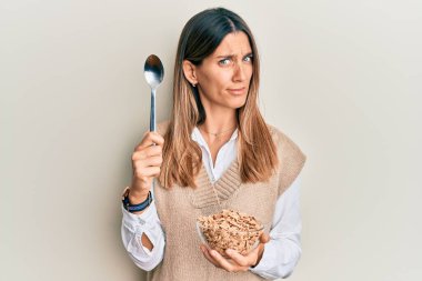 Esmer genç bir kadın sağlıklı tahıl gevrekleri yiyor. Kaşıksız ve kafası karışık bir ifade takınıyor. şüphe kavramı. 