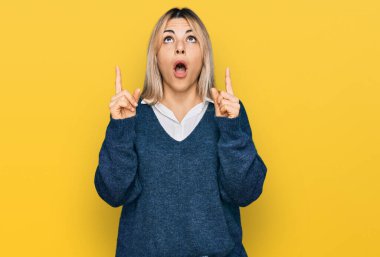 Gündelik giysiler giyen genç beyaz kadın şaşkın şaşkın yukarı bakıyor ve parmaklarıyla işaret ediyor ve kollarını kaldırıyor.. 