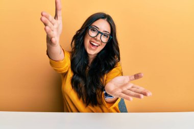 Güzel, esmer, gözlüklü ve gündelik giysileri olan genç bir kadın masaya oturmuş kameraya bakıyor ve sarılmak için kollarını açmış gülümsüyor. Mutluluğu kucaklayan neşeli bir ifade.. 