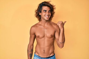Genç İspanyol adam gülen yüzüyle üstsüz duruyor başparmağıyla yan tarafa bakıyor ve işaret ediyor.. 