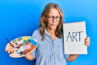 Boyacı paleti tutan orta yaşlı sarışın kadın şüpheci ve gergin, sorun yüzünden surat asıyor. negatif kişi. 