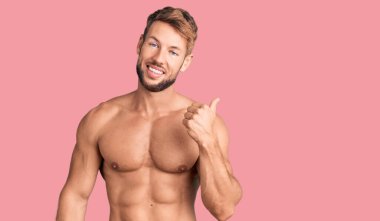 Genç, beyaz, tişörtsüz, gülen yüzlü bir adam başparmağıyla yan tarafa bakıyor ve işaret ediyor.. 