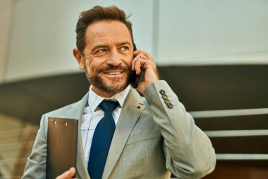 Orta yaşlı işadamı gülümsüyor. Şehirdeki akıllı telefondan konuşuyor..