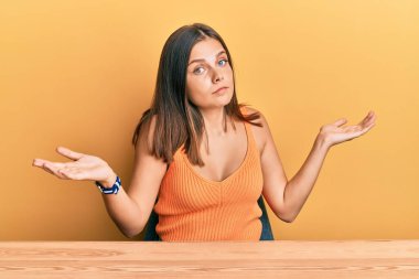 Gündelik giysiler giymiş genç beyaz bir kadın masanın üzerinde elleri ve kolları havada, kafası karışmış bir şekilde oturuyor. şüphe kavramı. 