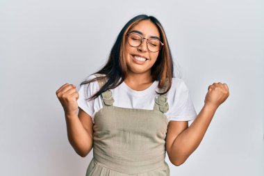 Gündelik kıyafet ve gözlük takan genç Latin kadın çok mutlu ve heyecanlı kolları havada, gülümseyen ve başarı için bağıran kazanan jestler yapıyor. kutlama konsepti. 