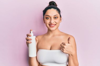 Güzel orta doğulu kadın elinde kozmetik nemlendirici krem, gülümsüyor mutlu ve pozitif, başparmağıyla mükemmel ve onay işareti yapıyor. 