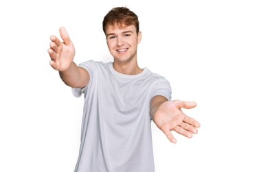 Beyaz bir tişört giyen genç beyaz bir adam kameraya sarılmak için kollarını açmış gülümsüyor. Mutluluğu kucaklayan neşeli bir ifade.. 