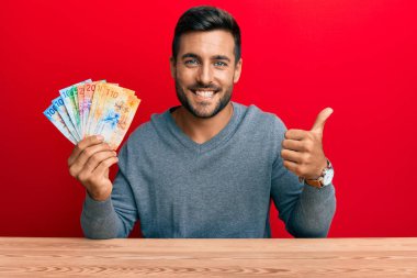 Yakışıklı İspanyol adam İsviçre frangı banknotları tutuyor mutlu ve olumlu gülümsüyor, başparmağıyla mükemmel ve onay işareti yapıyor. 
