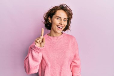 Kış süveteri giyen genç esmer kadın bir fikir veya soruyla gülümsüyor ve mutlu bir yüzle işaret ediyor. 
