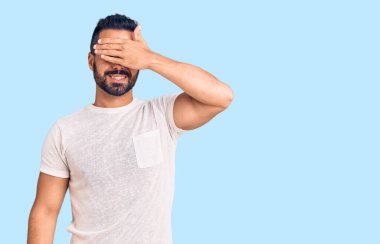 Gündelik kıyafetler giyen, gülümseyen ve gülümseyen genç İspanyol adam şaşırmak için gözlerini kapatıyor. kör kavram. 