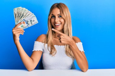 Dolarları tutan güzel sarışın genç kadın mutlu bir şekilde el ve parmağınla işaret ediyor. 