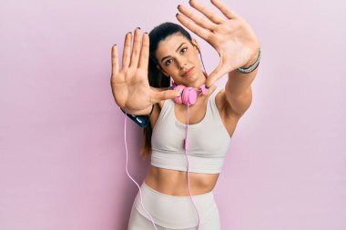 Spor kıyafetleri giyen, kulaklık takan, el ve parmaklarını kullanarak çerçeve yapan Latin kökenli genç bir kadın. 