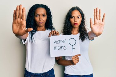 Afrika kökenli Amerikalı anne ve kız ellerinde kadın hakları pankartı tutuyorlar ve ciddi ve kendinden emin bir ifadeyle dur işareti yapıyorlar, savunma jesti. 