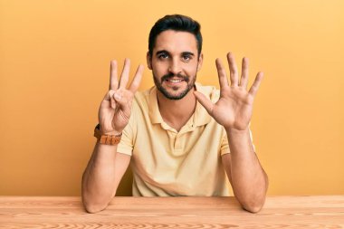 Latin asıllı genç adam, gündelik giysiler içinde masada oturuyor ve 8 numaralı parmaklarıyla işaret ediyor. Kendine güvenen ve mutlu bir şekilde.. 