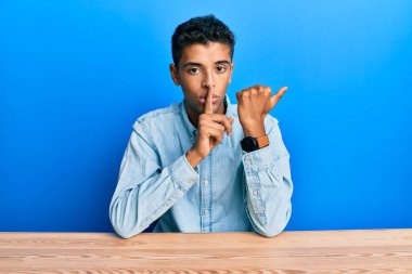 Genç, yakışıklı, Afro-Amerikalı, gündelik giysiler giymiş masada oturuyor ve parmakları yandan işaret eden dudaklarıyla sessiz olmasını istiyor. sessizlik ve gizli konsept. 