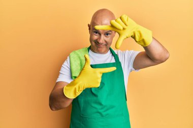 Orta yaşlı, kel, önlük takan, temizlik eldivenleri tutan, gülümseyen, elleri ve parmakları gülen yüzlü bir adam. yaratıcılık ve fotoğrafçılık kavramı. 