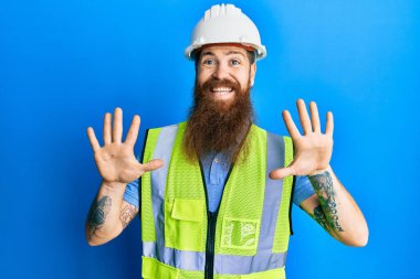 Uzun sakallı, güvenlik kaskı takan ve yansıtıcı ceketli kızıl saçlı adam on numaralı parmaklarını gösterirken kendinden emin ve mutlu bir şekilde gülümsüyor.. 
