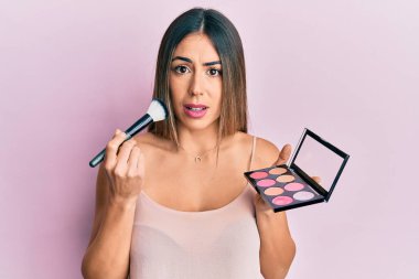 Makyaj fırçası tutan genç İspanyol kadın ve yüzü kızardı. şüphe kavramı. 