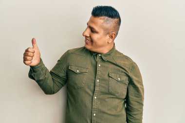 Gündelik kıyafetler giyen genç Latin adam gururlu görünüyor, gülümsüyor yan tarafa baş parmak işareti yapıyor. 