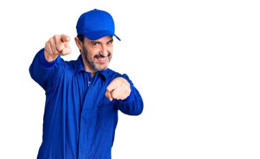 Orta yaşlı, tamirci üniforması giyen yakışıklı bir adam seni ve kamerayı parmaklarıyla göstererek mutlu ve mutlu gülümsüyor. 