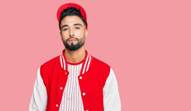 Sakallı, beyzbol forması giyen, yüzünde ciddi bir ifade olan genç bir adam. Basit ve doğal bir şekilde kameraya bakıyorsun.. 