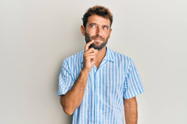 Sakallı, sıradan tişörtlü yakışıklı, şüpheye odaklanmış, çenesinde parmak olan ve kafasını kaldırıp merak eden bir adam. 