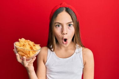 Güzel esmer küçük kız patates cipsi tutuyor. Korkmuş ve ağzı açık kalmış, şaşırmış ve şaşırmış. 
