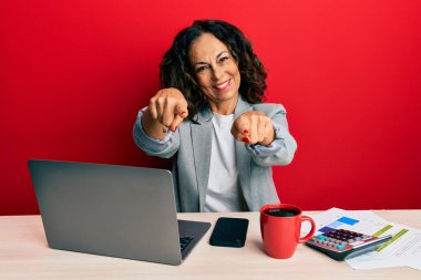 Ofiste çalışan orta yaşlı güzel bir kadın bir fincan kahve içiyor. Seni ve kamerayı işaret ediyor. 