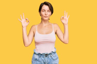 Kısa saçlı, kısa saçlı, yaz kıyafetleri giyen, gözleri kapalı gülümseyen ve parmaklarıyla meditasyon yapan genç esmer kadın. yoga konsepti. 