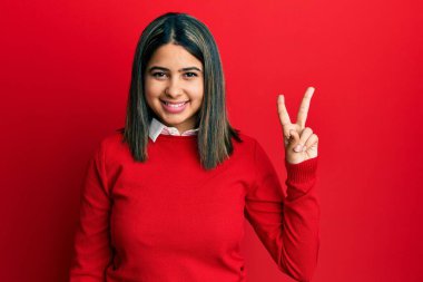 Gündelik kıyafetler giyen genç Latin kadın iki numarayı gösterip işaret ederken kendinden emin ve mutlu bir şekilde gülümsüyor.. 