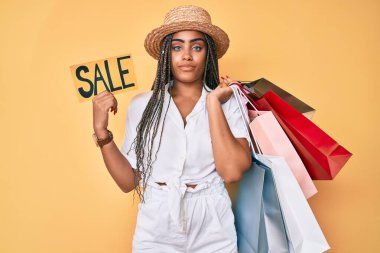 Saçları örgü örgülü, elinde alışveriş poşetleri ve satış afişi olan genç Afro-Amerikan kadın yüzünde ciddi bir ifadeyle rahatladı. Basit ve doğal bir şekilde kameraya bakıyorsun.. 