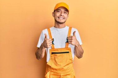 Tamirci üniforması giyen İspanyol genç adam el hareketleriyle olumlu jest yapıyor, gülümsüyor ve mutlu. Neşeli ifade ve kazanan jest. 