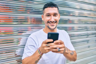 Duvara dayanmış akıllı telefon kullanan, mutlu gülümseyen genç İspanyol adam.