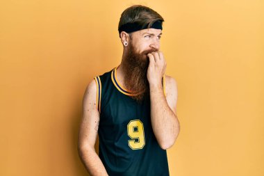Uzun sakallı, basketbol forması giyen, gergin ve elleri ağız yiyen kızıl saçlı bir adam. anksiyete. 