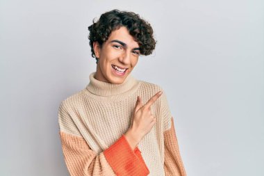 Hispanik genç adam, günlük kışlık süveter giyiyor, neşeli, yüzünde mutlu ve doğal bir ifadeyle, elleri ve parmaklarıyla yüzünü gösteren bir gülümseme var. 