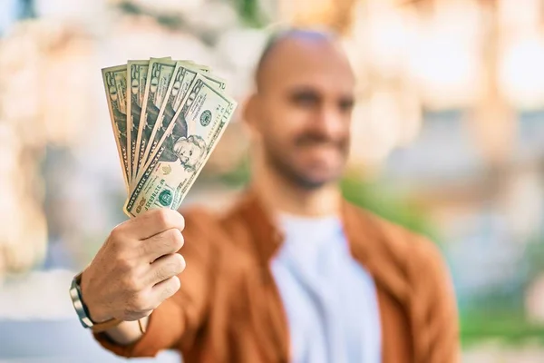 Young hispanic bald man smiling happy holding usa dollars at the city.