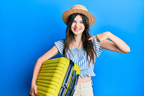 Young beautiful caucasian girl wearing summer dress and holding cabin bag pointing finger to one self smiling happy and proud