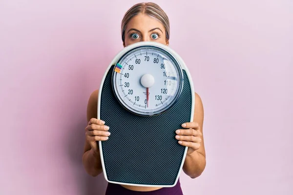 Beautiful blonde woman holding weight machine to balance weight loss making fish face with mouth and squinting eyes, crazy and comical.