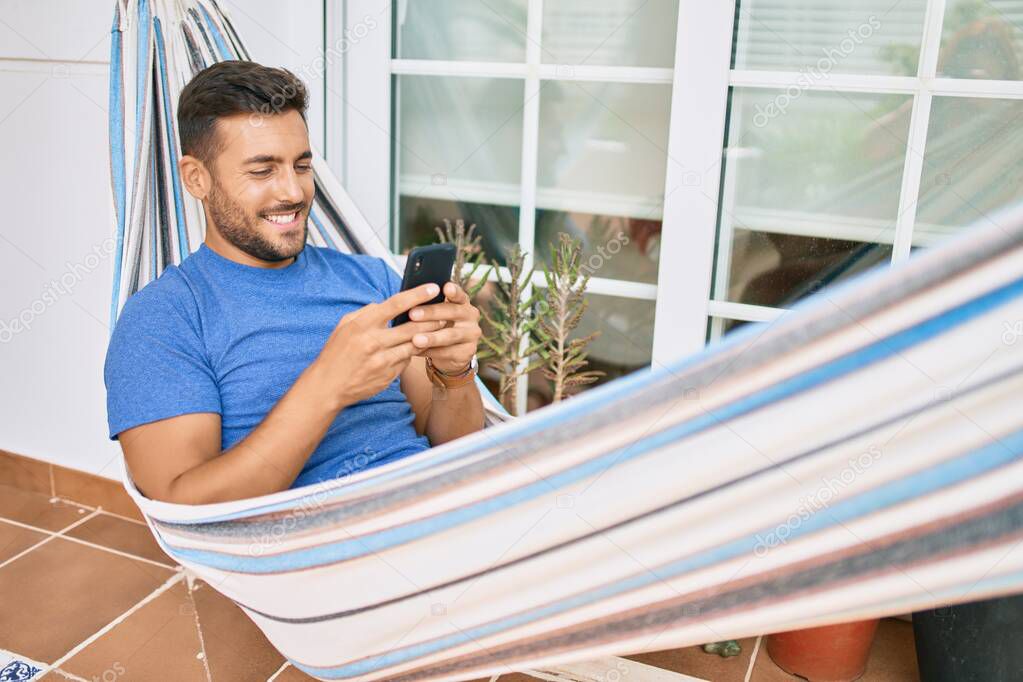 Young hispanic man relaxed using smartphone lying on the hammock at terrace.