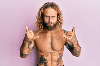 Sakallı, uzun saçlı, tişörtsüz, Shaka işareti yapan, şüpheci ve sinirli, sorun yüzünden surat asan yakışıklı bir adam. negatif kişi. 
