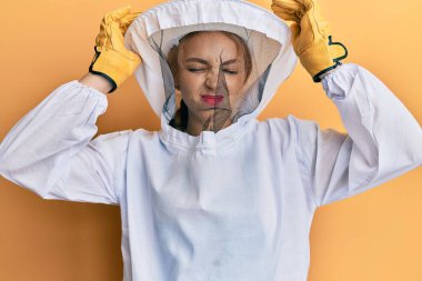 Güzel sarışın, beyaz kadın koruyucu arı üniforması giyiyor. Ağrı ve migren yüzünden çaresiz ve stresli baş ağrısı çekiyor. Eller başının üstünde.. 