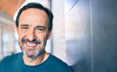 Orta yaşlı, sakallı, mutlu, açık havada duvara yaslanmış bir adam.