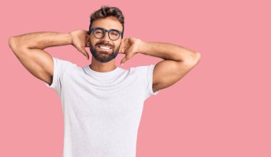 Genç İspanyol adam günlük kıyafetler giyiyor ve gözlük takıyor rahatlıyor ve esniyor, kolları ve elleri başının ve boynunun arkasında mutlu bir şekilde gülümsüyor. 