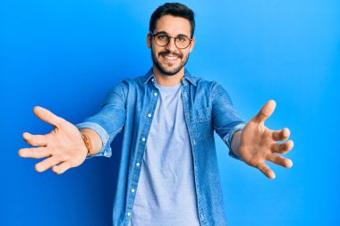 Gündelik giysiler ve gözlük takmış genç İspanyol adam kameraya bakıp sarılmak için kollarını açmış gülümsüyor. Mutluluğu kucaklayan neşeli bir ifade.. 