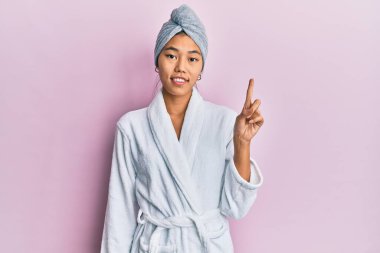 Duş havlusu şapkası ve bornoz takan genç Çinli kadın bir numaralı parmağını gösterip işaret ederken kendinden emin ve mutlu bir şekilde gülümsüyor.. 