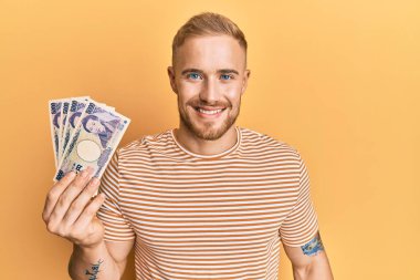 Elinde Japon Yen banknotları tutan genç beyaz bir adam. Olumlu ve mutlu görünüyor. Kendine güvenen bir gülümsemeyle dişlerini gösteriyor. 