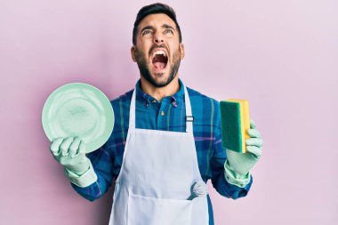 Önlük giymiş İspanyol bir genç bulaşık yıkıyor kızgın ve kızgın çığlıklar atıyor öfkeyle bağırıyor öfkeyle yukarı bakıyor. 